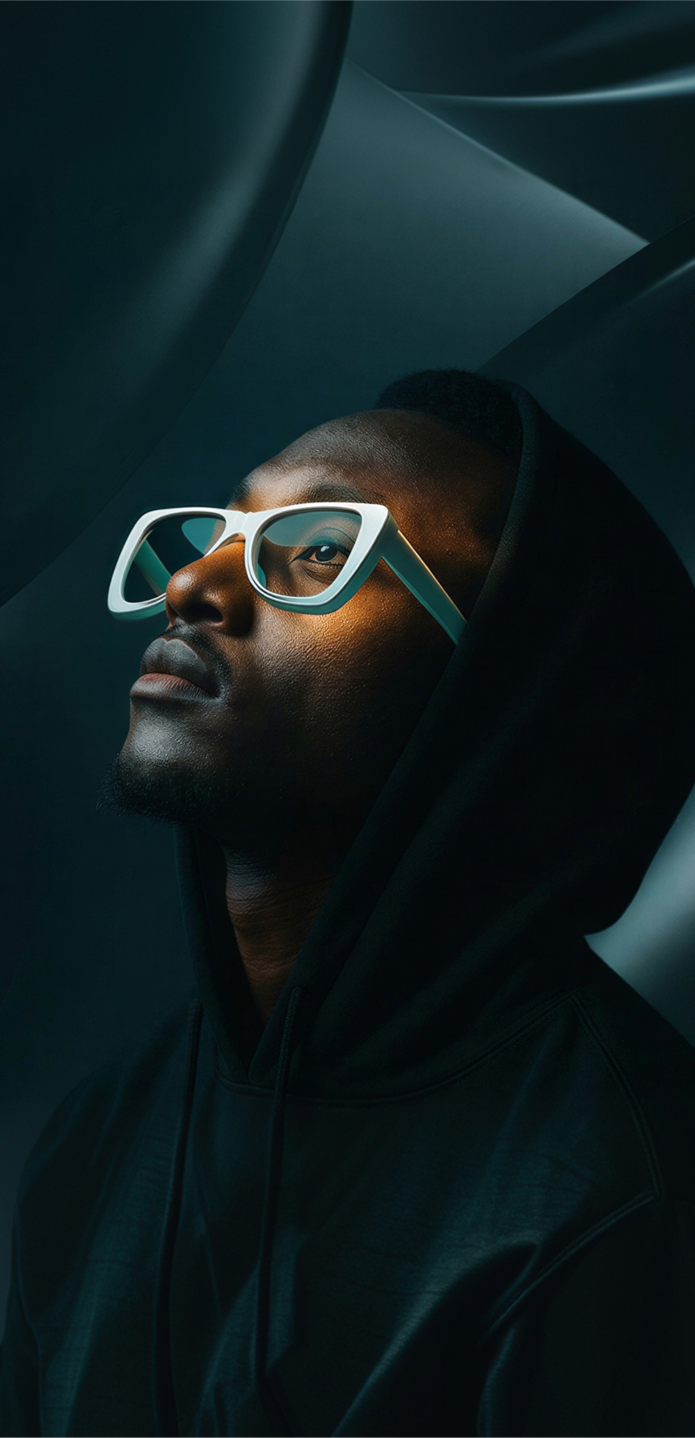 Portrait of a man in white-framed glasses looking upward, wearing a dark hoodie.