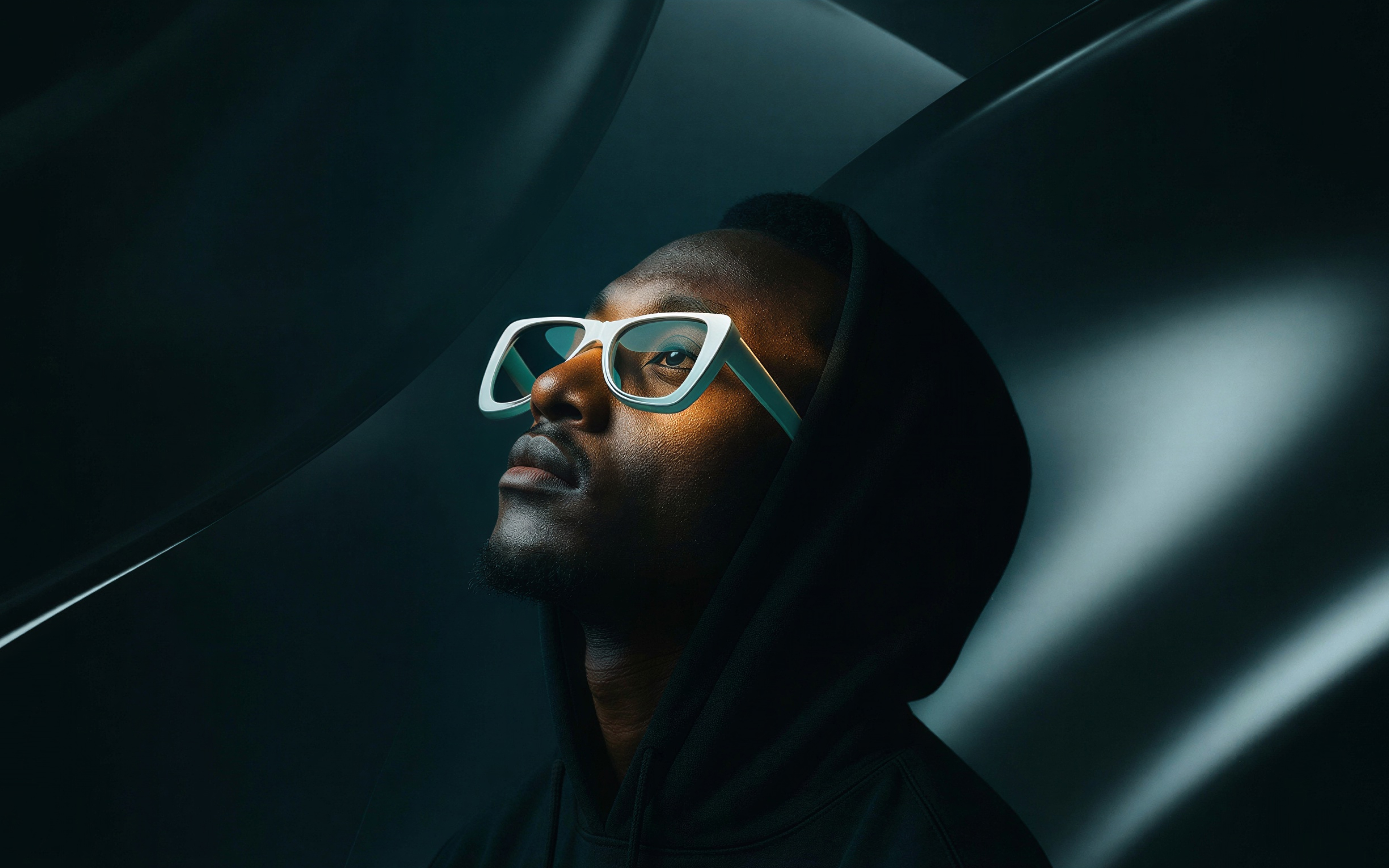 Portrait of a man in white-framed glasses looking upward, wearing a dark hoodie.